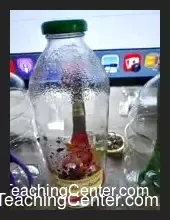 Close-up of a pair of metal scissors mysteriously standing upright inside a clear glass bottle, with a small silver padlock secured through the finger holes of the scissors, suggesting an impossible feat of magic. The bottle sits on a dark, polished surface, highlighting the illusion.